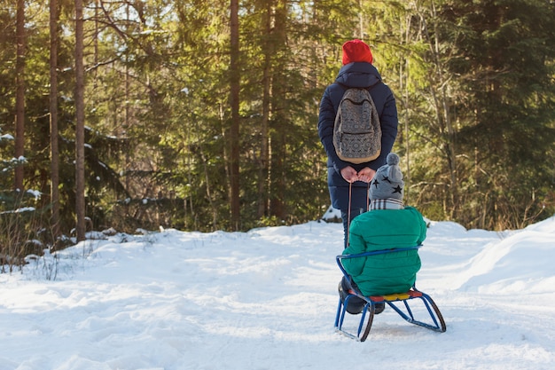 Mamá lleva a su hijo en un trineo por el bosque de coníferas cubierto de nieve.