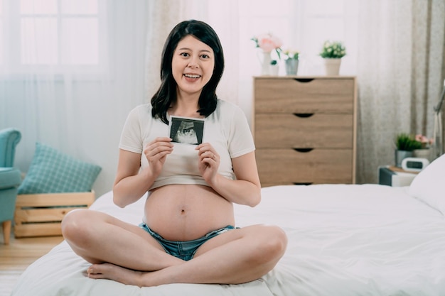 mamá de maternidad mostrando una cámara sonriendo mientras sostenía una foto de ultrasonido sentada en la cama. mujer china asiática embarazada alegre relajarse en el dormitorio después de volver a casa del hospital de examen de parto.