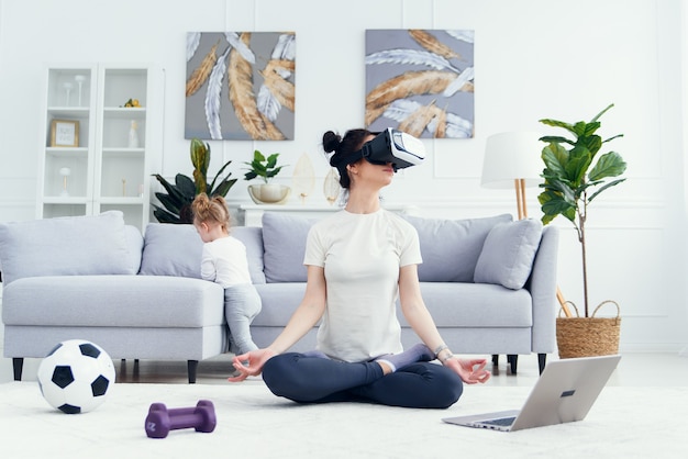 Mamá meditando en posición de loto yoga con su hija en casa.