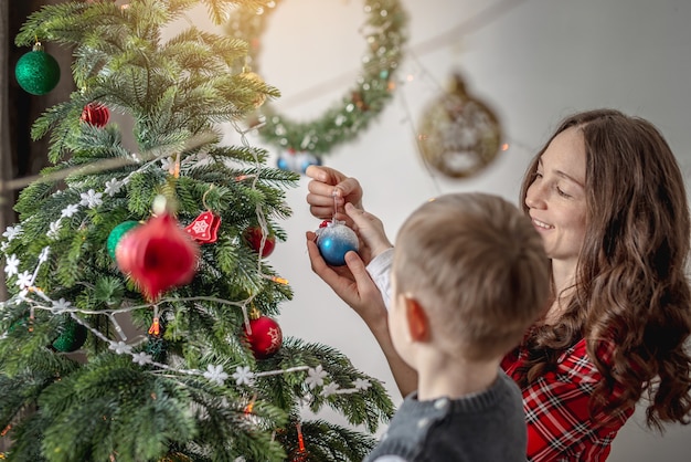 Mama mit ihrem Kind schmückt zusammen den Weihnachtsbaum mit Spielzeug und Bällen. Konzept der Familienvorbereitung für das neue Jahr, gemütliche Atmosphäre und festliche Stimmung.