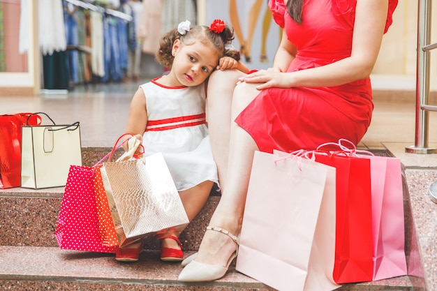 Mama mit kleiner Tochter in Kleidern sitzt mit bunten Taschen auf den Stufen im Einkaufszentrum