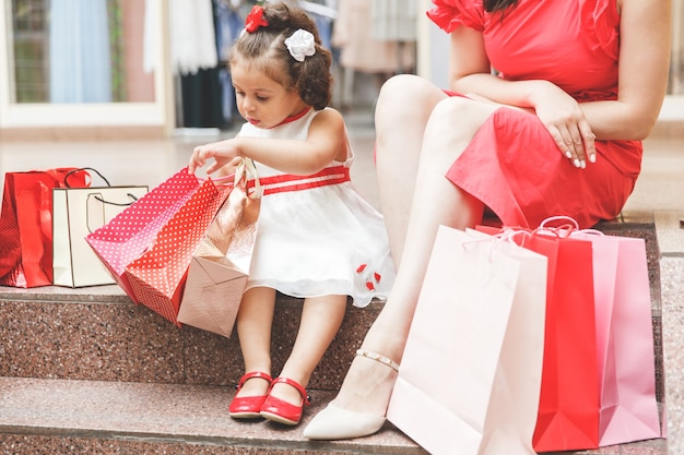 Mama mit kleiner Tochter in Kleidern sitzt mit bunten Taschen auf den Stufen im Einkaufszentrum