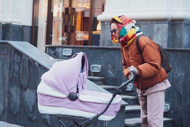 Mamá moderna de moda con un cochecito en un paseo por la ciudad, otoño. Mamá conduce un cochecito por las calles, de cerca.