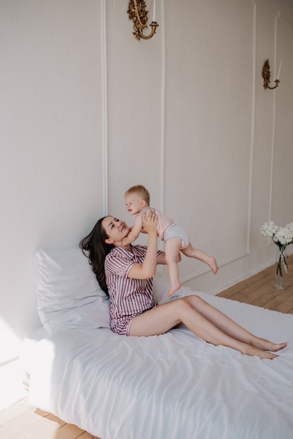 Mamá morena hermosa en pijama juega con su hija en la cama en la habitación