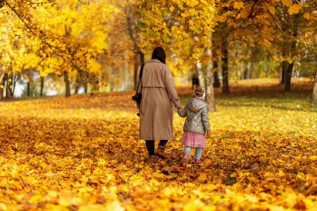 Mamá mujer con hija niño en ropa de moda caminando sobre follaje otoñal amarillo en el parque de otoño