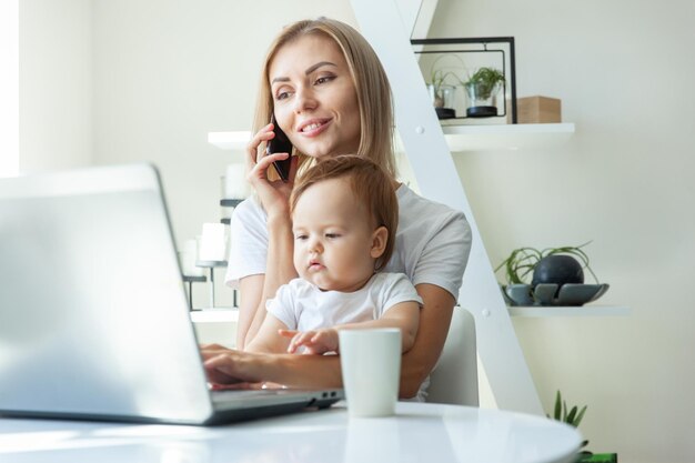 Mamá de negocios con su hija pequeña en los brazos usa una computadora portátil y habla por teléfono mientras está sentada en una mesa en casa