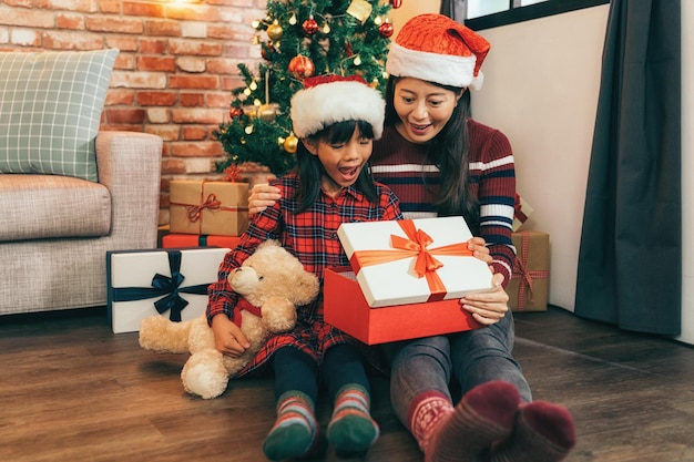 Foto mamá y una niña linda con su osito de peluche favorito sentados en el suelo de madera en la sala de estar. familia abriendo regalo de navidad juntos. celebrando con alegría el concepto de navidad en casa.