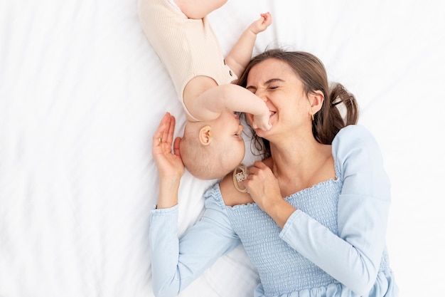 Mamá con una niña recién nacida en un traje de algodón en la cama en casa abrazándolo y besándolo, feliz maternidad o familia