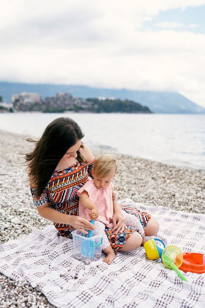 Mamá y niña se sientan en una colcha en una playa de guijarros y juegan con juguetes