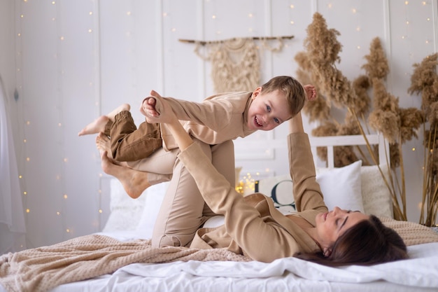 Mamá y un niño pequeño juegan a acurrucarse y hacen un avión en casa en una cama en tonos marrones naturales una familia feliz con un niño pequeño se divierten juntos