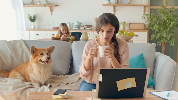 Mamá ocupada tratando de trabajar de forma remota desde casa. Al fondo, su hija está sentada en un escritorio, mientras que su madre trabaja con una computadora portátil y tiene una conversación de negocios por teléfono.