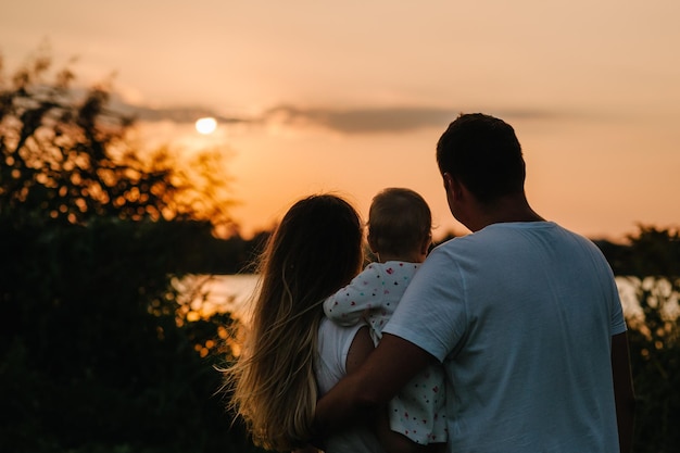 Mamá papá detiene a su hija y mira la puesta de sol Retrato de una familia joven en la naturaleza de vacaciones al aire libre El concepto de vacaciones familiares