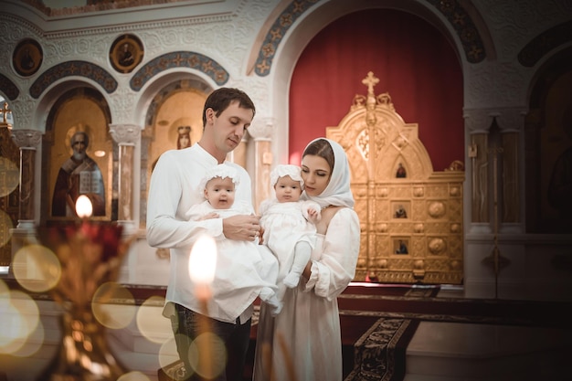 Mamá papá de familia con niños pequeños gemelos en un templo o iglesia rezando cerca de un ícono y velas o vino a adorar en la iglesia ortodoxa rusa el bautismo de un bebé