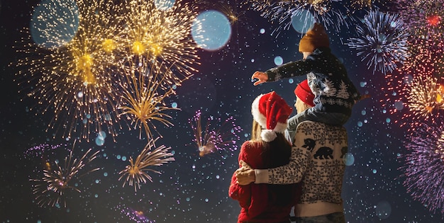 Mamá y papá con su hijo sobre los hombros con ropa abrigada y un gorro de Papá Noel La familia celebra el año nuevo mirando los fuegos artificiales afuera