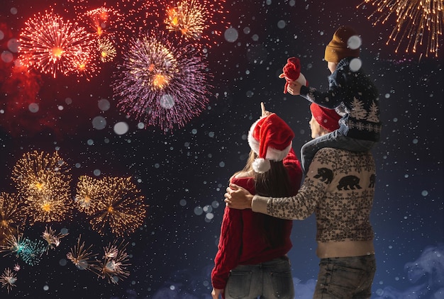 Mamá y papá con su hijo sobre los hombros con ropa abrigada y un gorro de Papá Noel La familia celebra el año nuevo mirando los fuegos artificiales afuera