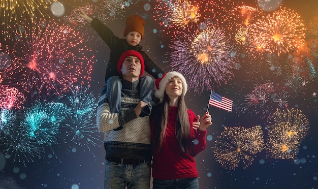Foto mamá y papá con su hijo sobre los hombros con ropa abrigada y un gorro de papá noel la familia celebra el año nuevo mirando los fuegos artificiales afuera