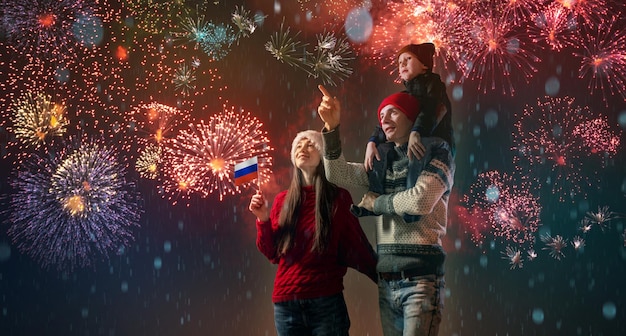 Mamá y papá con su hijo sobre los hombros con ropa abrigada y un gorro de Papá Noel La familia celebra el año nuevo mirando los fuegos artificiales afuera