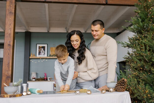 Mama, Papa und kleiner Sohn schneiden Formen aus Teig für Lebkuchen oder Lebkuchenhaus aus. Vorbereitung auf Weihnachten