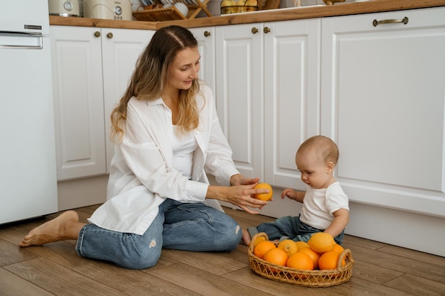 mamá en el piso de la cocina con un bebé y una canasta de frutas le da una naranja