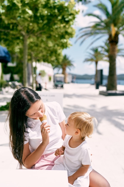 Mama probiert Eis vor einem kleinen Mädchen, das auf ihrem Schoß in einem Café am Strand sitzt