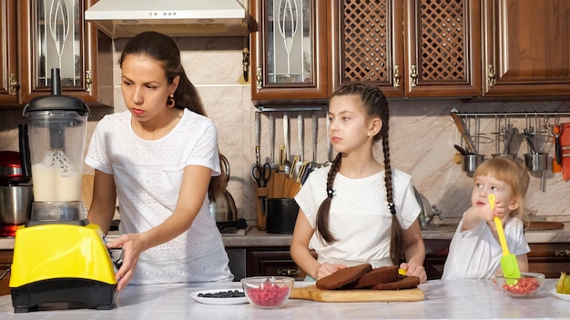 Mama probiert und kocht Sahne für Kuchen mit einem Mixer mit ihren beiden Töchtern in der Küche zu Hause, Mädchen betrachten den Prozess mit Interesse, kleines Mädchen isst Beeren mit kulinarischem Spatel.