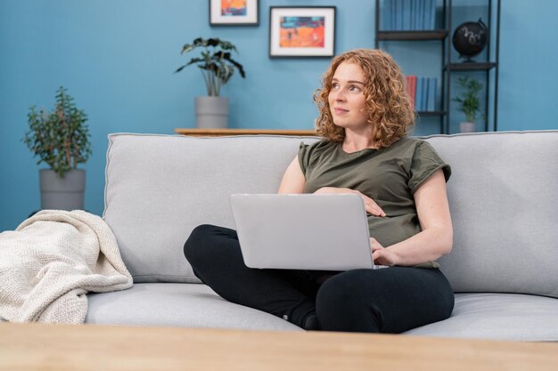 Mamá real pensativa hermosa mujer embarazada sentada en un sofá en casa con una computadora portátil en sus manos