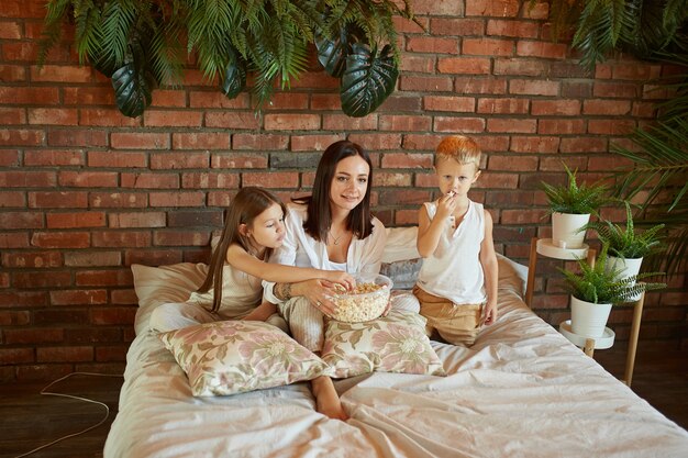 Mamá se sienta en la cama con su hijo y su hija y mira una película.