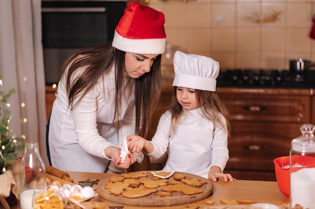 Mamá con sombrero de Papá Noel con su pequeña hija decora pan de jengibre con glaseado Navidad y año nuevo