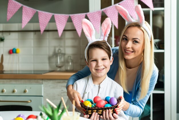 Mamá sonriente con el hijo que presenta sosteniendo los huevos de Pascua en oídos de conejo.