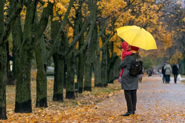 Mamá sostiene a un niño pequeño en sus brazos mientras está de pie bajo un paraguas amarillo en un parque de otoño contra un fondo de follaje amarillo