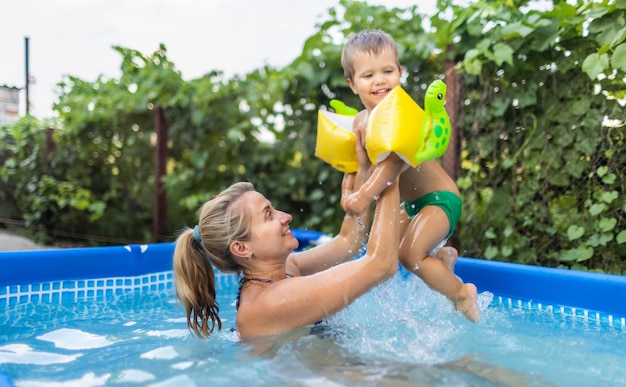 Mama spielt mit einem nackten Baby in Ärmeln im Pool vor dem Hintergrund eines Sommersonnenuntergangs