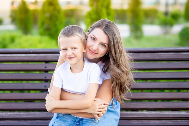 Mamá y su hijo en el parque en un banco en verano se divierten abrazándose riendo y jugando