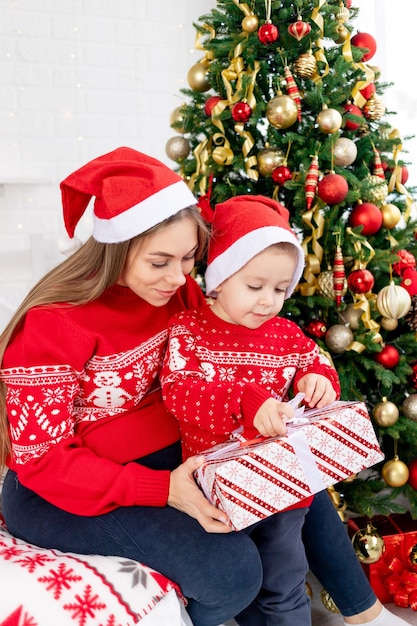 Mamá y su hijo con un suéter rojo y sombreros dan regalos bajo el árbol de Navidad en casa y se regocijan en el año nuevo y la Navidad.