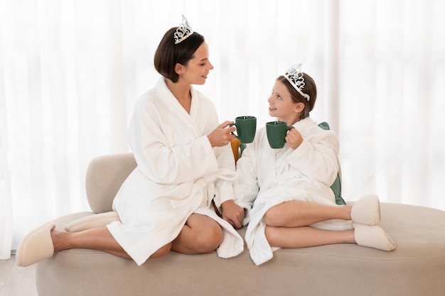 Foto mamá y su pequeña hija vistiendo albornoces y coronas de juguete relajándose en el sofá