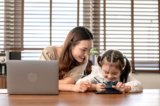 Mamá trabajando y enseñando a su hija para el aprendizaje en línea en casa. Estilo de vida de unión familiar y nueva normalidad después de Covid-19.