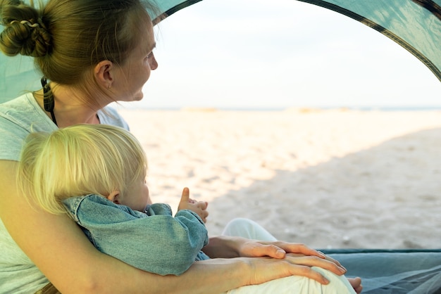 Mama umarmt das Baby, während sie im Touristenzelt sitzt. Meer- und Strandblick vom Campingzelt.