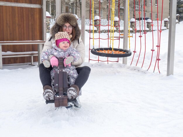 Mama und Baby reiten im Winter an einem sonnigen Tag auf einer Schaukel auf dem Spielplatz