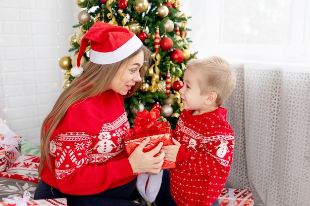 Mama und ihr kleiner Sohn in einem roten Pullover und Mützen machen zu Hause Geschenke unter dem Weihnachtsbaum und freuen sich auf das neue Jahr und Weihnachten