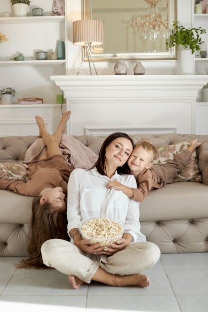 Mama und Kinder essen an einem freien Tag zu Hause Popcorn. Eine Frau, ein Junge und ein Mädchen entspannen sich auf der Couch und umarmen sich