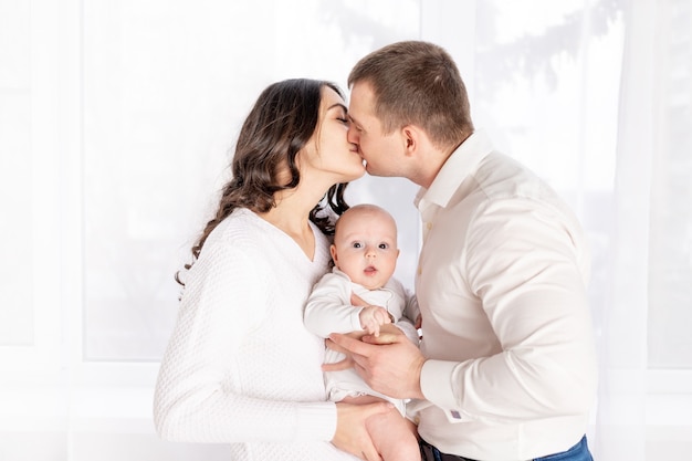 Mama und Papa küssen sich und halten ein neugeborenes Baby zu Hause am Fenster, das Konzept einer glücklich liebenden Familie, Lebensstil