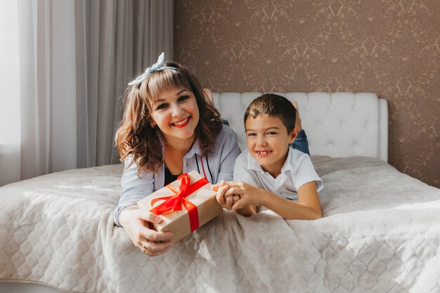 Mama und Sohn liegen auf dem Bett. Kind gibt seiner Mutter ein Geschenk zum Muttertag.