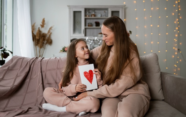 Mama und Tochter sitzen auf dem Sofa im Zimmer und halten eine Karte mit Herz