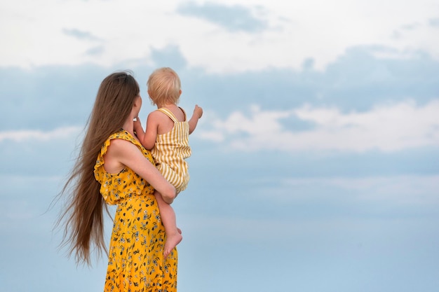 Mamá con vestido brillante y cabello largo sostiene al bebé y mira el mar Concepto de maternidad Vacaciones con niños