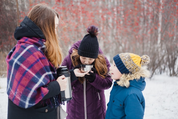 Mamá vierte té caliente o bebida del termo para su hija y su hijo, paseo de invierno, invierno