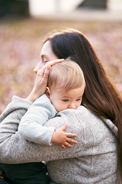 Mamãe abraça o bebê junto ao peito, sentada na folhagem no parque, close-up