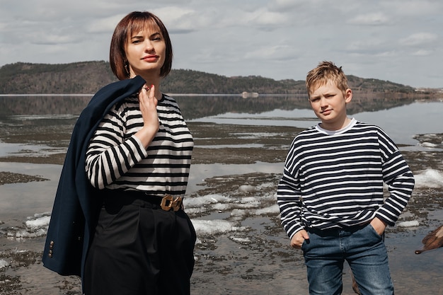 Mamãe abraça o filho na margem do rio. Uma mãe bonita e moderna com um filho feliz em coletes listrados.