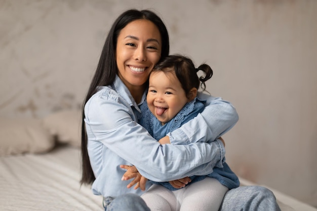Mamãe asiática abraçando bebê enquanto bebê mostra a língua dentro de casa