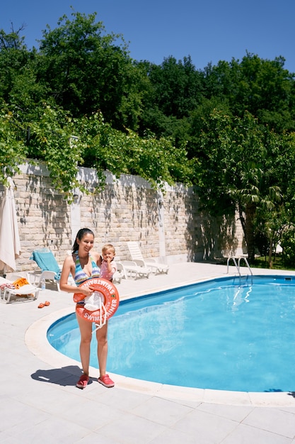 Mamãe está à beira da piscina com uma menina e um anel inflável nos braços