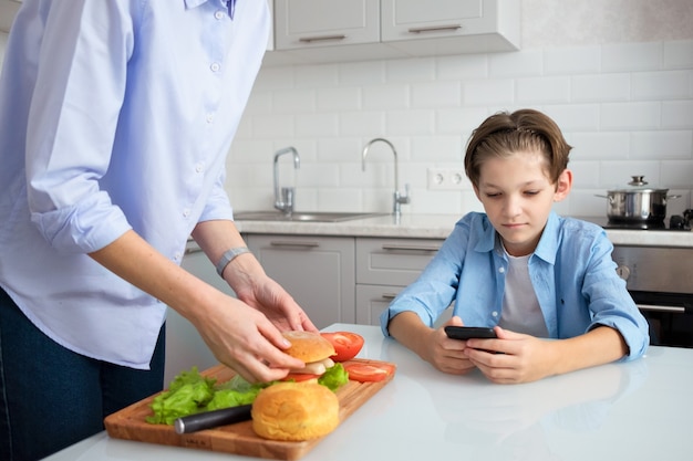 Mamãe está na cozinha preparando um hambúrguer caseiro para seu filho adolescente. Um menino de 11 anos está sentado à mesa da cozinha e olhando para o celular.
