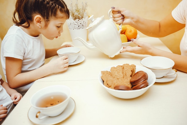 Mamãe jovem e bonita na cozinha com seus filhos bebendo chá com biscoitos